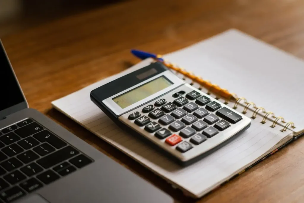 A calculator placed on a notebook with a pen and a laptop, symbolizing budgeting, financial planning, and expense tracking.